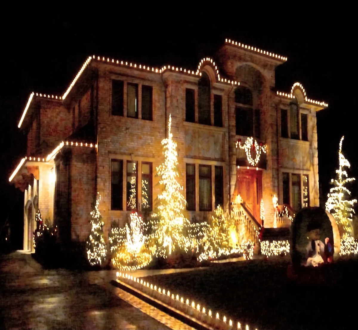 Brick home with lit rooflines and glowing cone trees across the front lawn