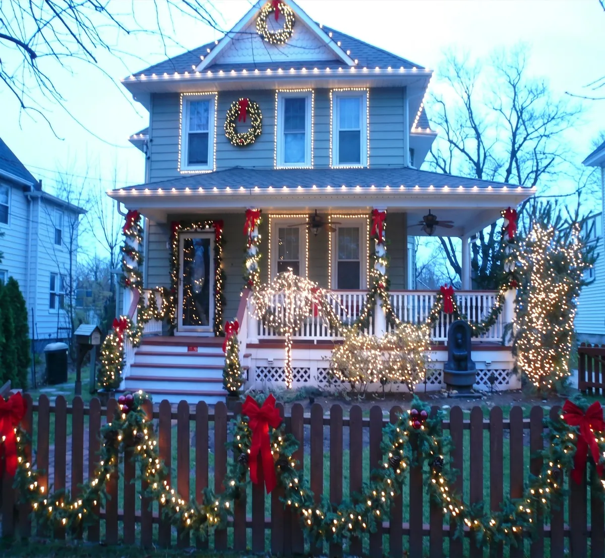 Front porch railings dressed in garland and red bows, with lit trees in the yard